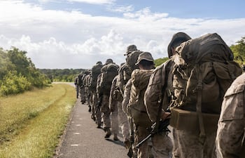 Grupo de soldados en uniforme camuflado con mochilas, avanzando sobre un camino flanqueado por vegetación densa.