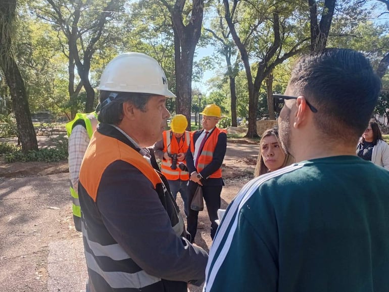 Hombre con casco blanco y chaleco naranja habla, mientras una mujer con cabello lacio escucha atentamente rodeada de personas con chalecos reflectantes.