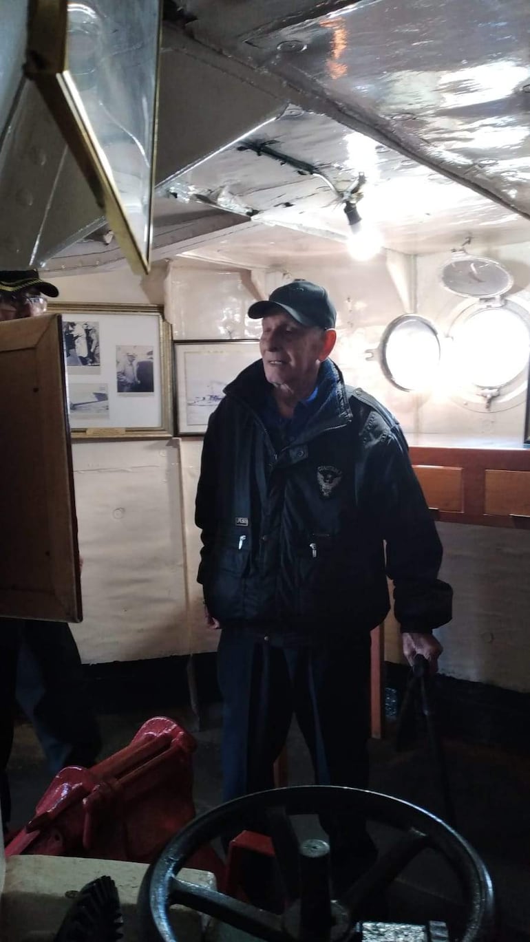 Sixto Sosa Ramírez, vestido con abrigo negro y gorra, habla con un marinero de uniforme en interior oscuro de un barco.