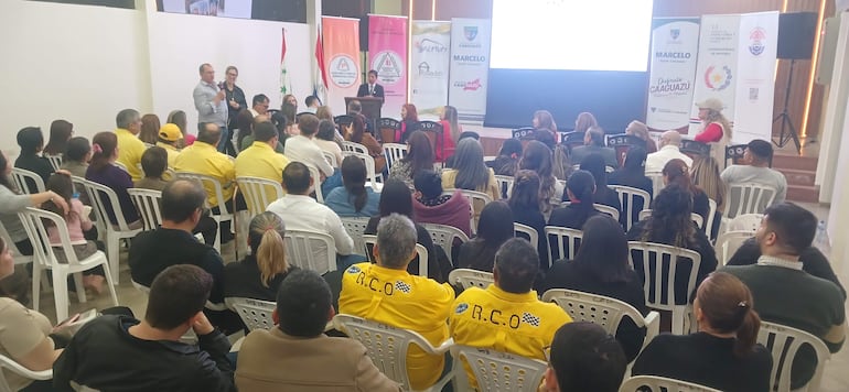 Panelistas discuten propuestas mientras asistentes en camisetas amarillas escuchan atentamente en un salón decorado formalmente.