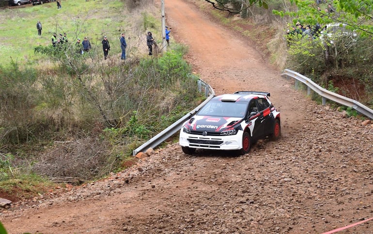 Volkswagen negro y blanco, detalles rojos en movimiento sobre camino de tierra; espectadores entre vegetación observan atentos.