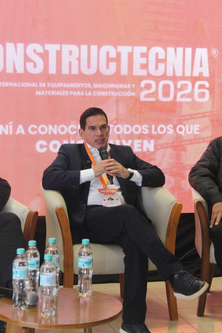 Juan Carlos Baruja en traje oscuro, sosteniendo un micrófono mientras dos hombres en sillas lo escuchan atentamente en un ambiente formal.