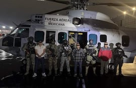 Seis hombres, entre ellos dos sin armas, posan frente a un helicóptero militar, iluminado en la oscuridad de la noche.