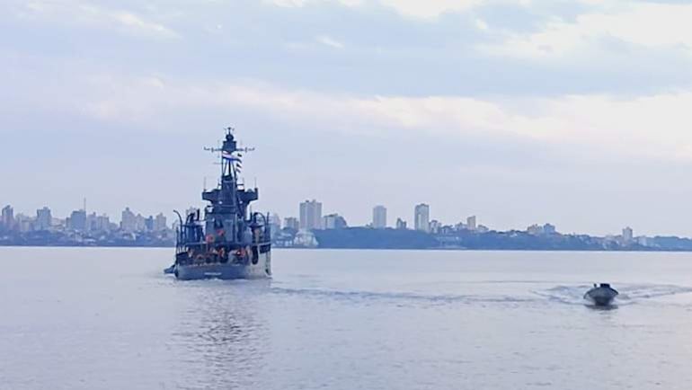 Buque naval Cañonero Paraguay C-1 navegando en agua cercana a una lancha motorizada, con edificios urbanos al fondo.
