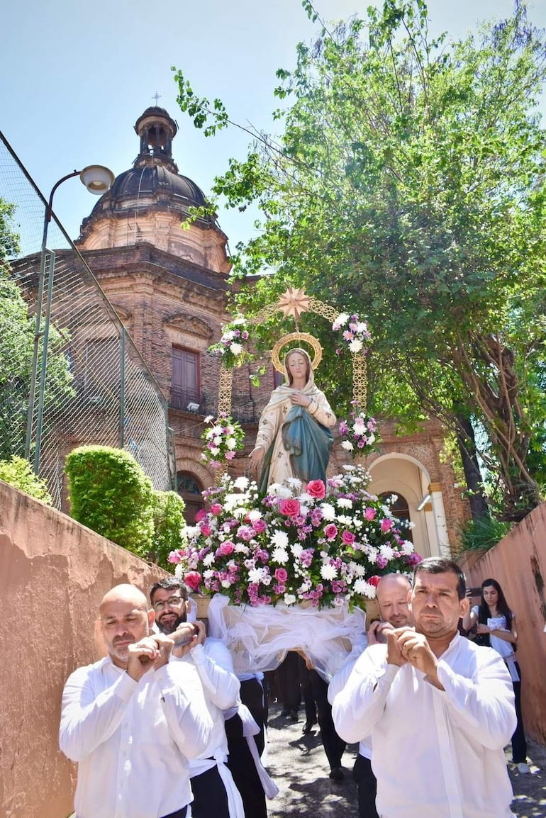 Cuatro hombres con camisas blancas llevan una imagen de la Virgen María adornada con flores, frente a un edificio antiguo.