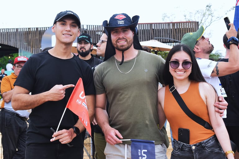 Tres personas sonrientes en primer plano; una con camiseta negra y bandera, otra con sombrero y camiseta verde, y una más con blusa naranja y gafas de sol.