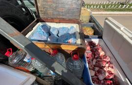 Recipientes con alimentos y agua dispuestos en transporte, incluyendo cajas de madera y hielera blanca, en un entorno exterior.