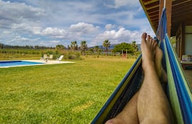 Una persona descansa con los pies descalzos en una hamaca paraguaya, en una quinta al costado de una pileta.