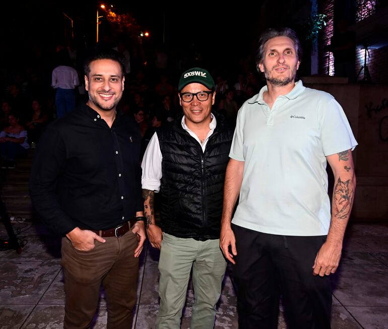 Tres hombres de pie: uno en camisa negra sonríe, otro con chaleco negro y gorra verde SXSW, y un tercero con camiseta clara y tatuajes.