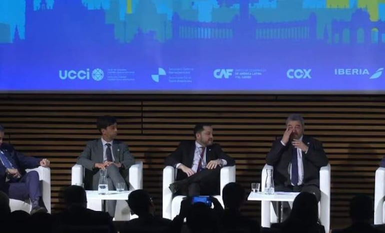 Luis Bello, con expresión triste, discute en panel con otros hombres en traje en un ambiente formal.