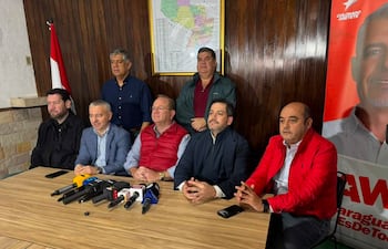 Camilo Perez en chaqueta roja, rodeado de hombres en camisas blancas y grises, en salón con bandera paraguaya y micrófonos.