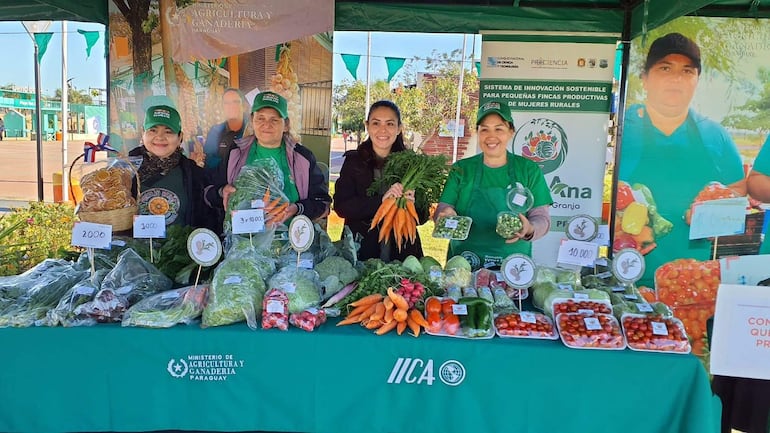 Cuatro mujeres sonrientes en un mercado agrícola, sosteniendo productos frescos, algunas vestidas con camisetas verdes y chaqueta morada.