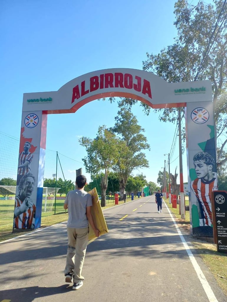 Hombre con camiseta gris y pantalones claros camina sosteniendo una caja cerca del arco 'ALBIRROJA' en ambiente deportivo.