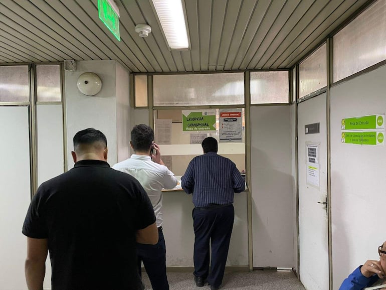 Cuatro personas esperan en oficina, un hombre en camisa de rayas realiza un trámite ante una ventanilla.