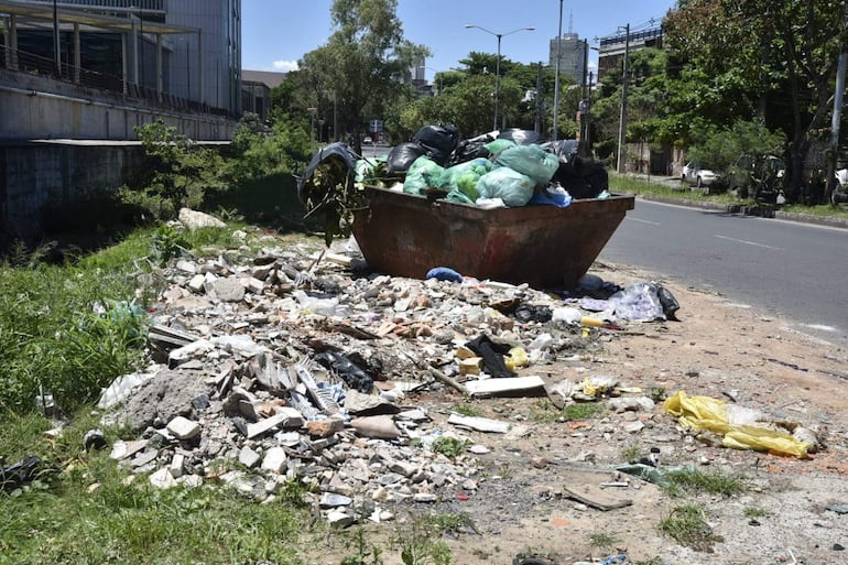 Contenedor de basura lleno de bolsas de diferentes colores y escombros en un ambiente urbano abandonado.