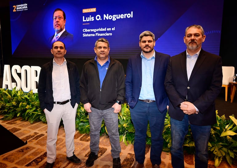 Cuatro hombres de pie en traje formal en evento de ciberseguridad, con cartel de Luis O. Noguerol al fondo.