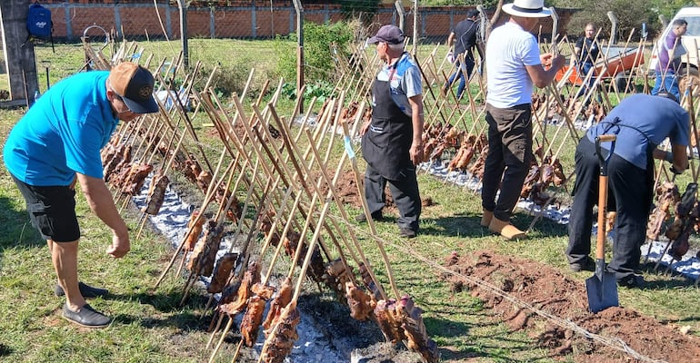 Hombres en ambiente rural, uno ajusta carne en la parrilla y otro observa con delantal negro, rodeados de césped y valla.