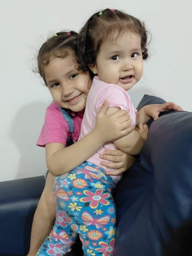 Dos niñas, una con camiseta rosa y overol azul, y otra con camiseta rosa y leggings floreados, se abrazan sonriendo en un sofá azul.
