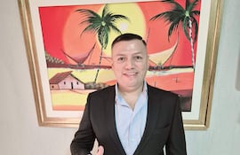 Hombre sonriente en traje negro y camisa azul claro, levantando el pulgar en un entorno con pintura tropical colorida.