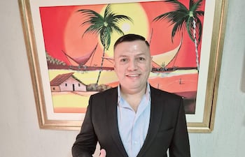 Hombre sonriente en traje negro y camisa azul claro, levantando el pulgar en un entorno con pintura tropical colorida.