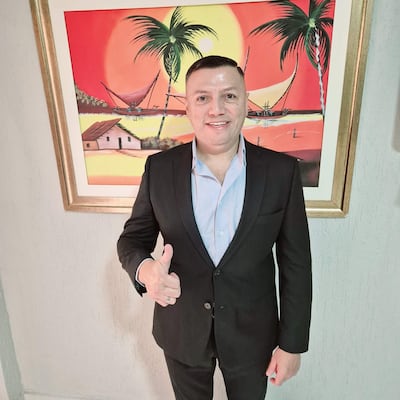 Hombre sonriente en traje negro y camisa azul claro, levantando el pulgar en un entorno con pintura tropical colorida.