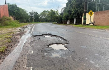 Carretera en mal estado con baches llenos de agua, rodeada de césped y árboles. No hay personas presentes.