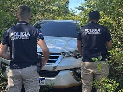 Dos agentes de policía de espaldas, vestidos con camisetas oscuras, observan una SUV blanca cubierta de maleza en un entorno natural.