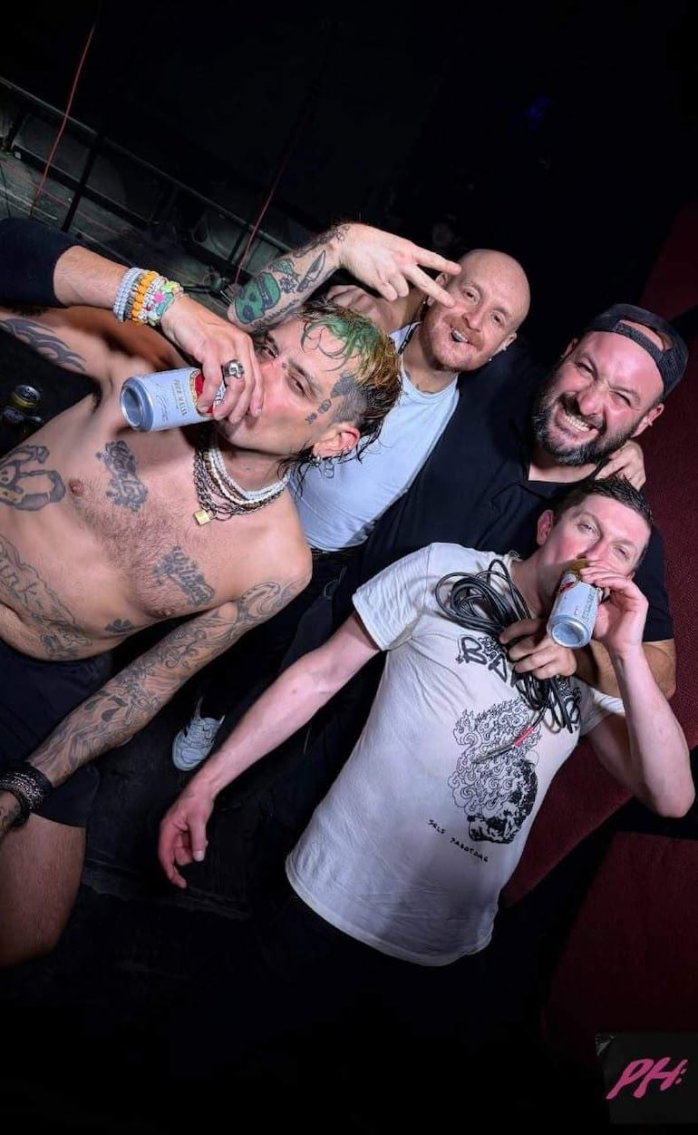 Cuatro hombres sonrientes en un bar, con tatuajes y latas de bebida, celebrando entre risas en un ambiente urbano.