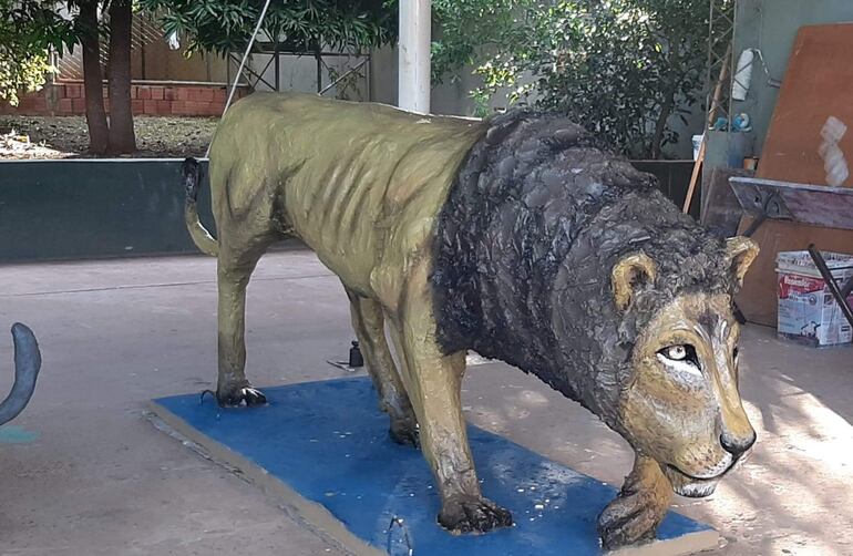 Escultura de león en tonos amarillos y marrones sobre base azul, rodeada de vegetación en un ambiente iluminado.
