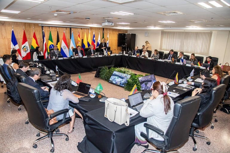 Reunión formal con 15 personas en trajes, algunos con laptops, en un salón con banderas de diferentes países.