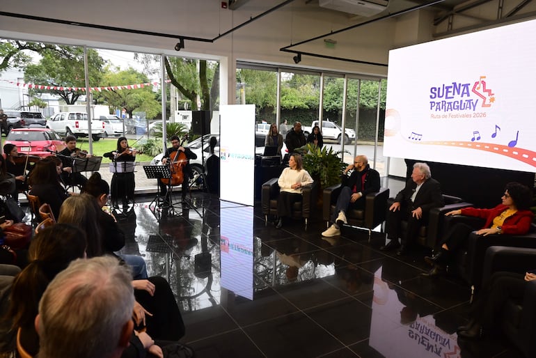 Panel de cuatro personas sentadas discutiendo, mientras un grupo musical toca instrumentos de cuerda en un ambiente moderno y colorido.