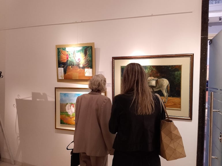 Dos mujeres en galería de arte, una con cabello canoso mirando un cuadro, la otra con cabello largo y bolsa de mano.