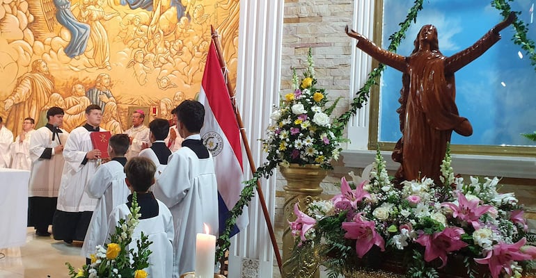 Jóvenes en túnicas blancas y diácono con libro rojo en ritual solemne, fondo con bandera paraguaya y altar decorado.