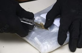 Manos enguantadas abren paquete plateado que parece contener droga en un ambiente interior difuso.