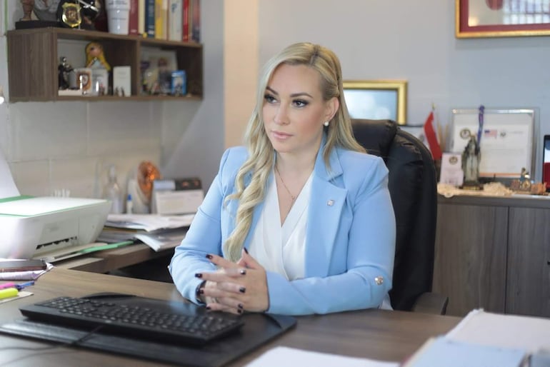 Vivian Coronel Peralta, con saco azul claro, observa atentamente en su oficina, rodeada de documentos y objetos de escritorio.