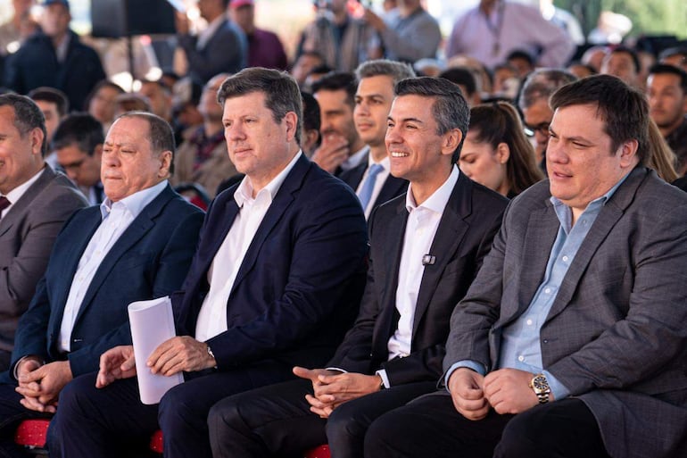 Cinco hombres en trajes formales, algunos sonrientes y otros serios, participan en un acto público al aire libre.