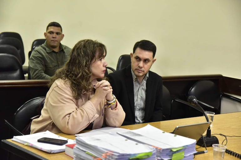 Alba Zaracho habla de pie, mientras Hernán Rivas escucha atento, con camisa de rayas y saco negro, en ambiente judicial.