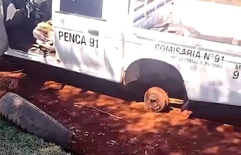Camioneta blanca de la Comisaría N° 91 aparcada, con una rueda desinflada, en un terreno de tierra con vegetación al fondo.