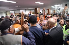 Grupo de funcionarios mostrando gestos intensos y organizados en un semicírculo, algunos levantan los brazos en un ambiente cerrado.