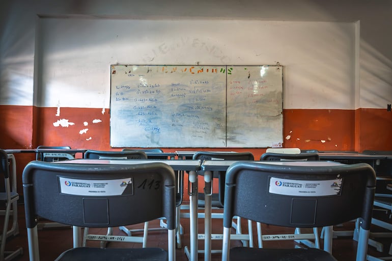 Aula vacía con bancos modernos y pizarra deteriorada, esperando la llegada de los estudiantes.