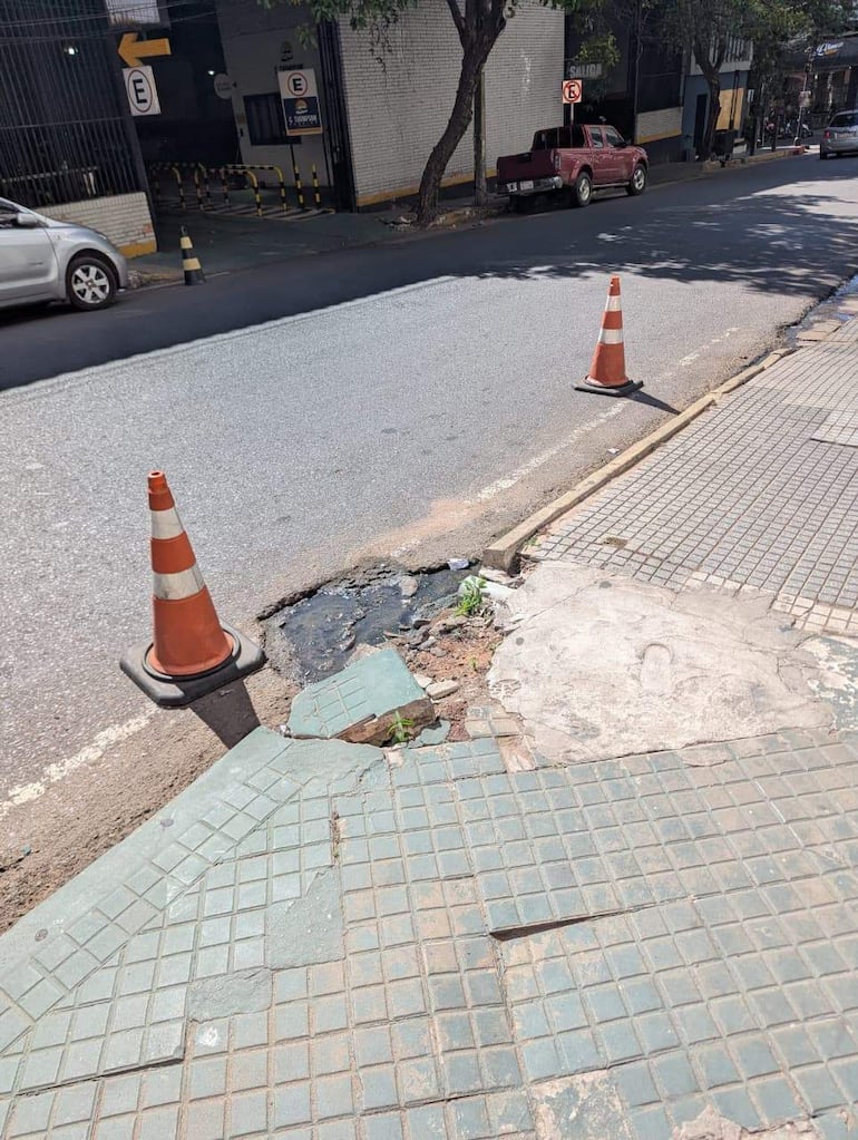 Pavimento roto y bache con agua en la esquina del MOPC; conos de tráfico marcan el área afectada.
