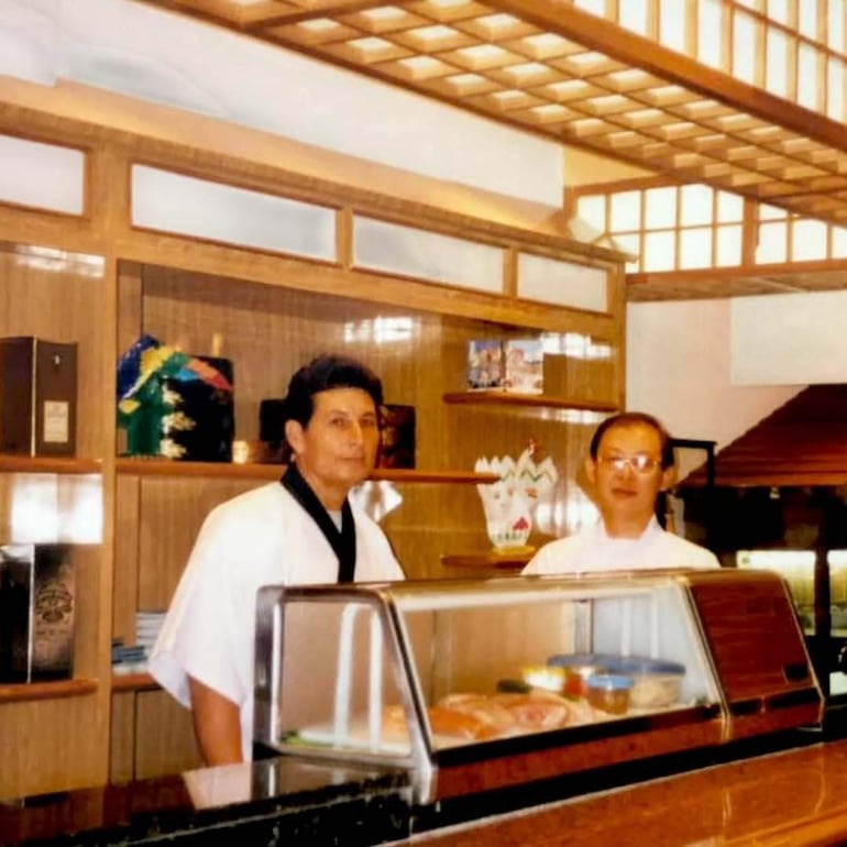 Diosnel Miranda en camisa blanca y delantal oscuro, junto a otro hombre, ambos de pie detrás de un mostrador en restaurante japonés.