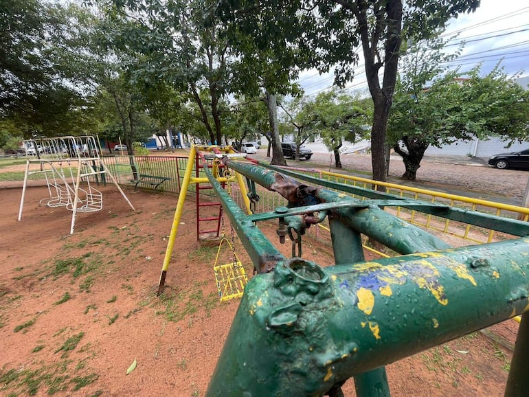 Estructuras de juegos metálicas oxidadas y descascaradas en un parque infantil sin niños presentes, rodeado de árboles.