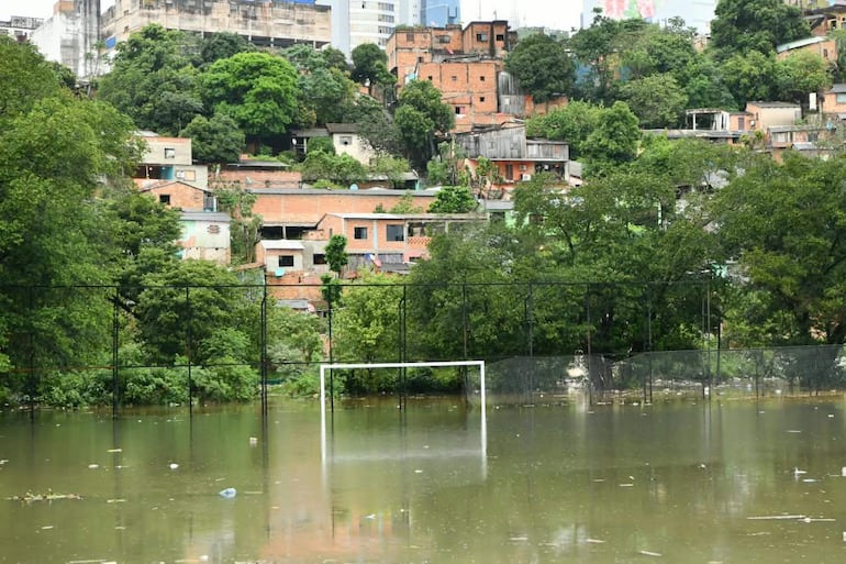 Campo de fútbol inundado, con agua cubriendo el área de juego y viviendas de ladrillo en colina al fondo.