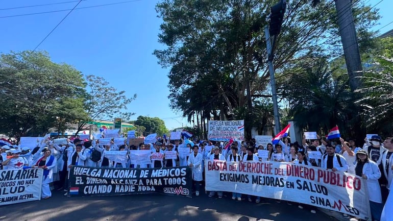 Grupo de médicos en batas blancas sostienen pancartas en protesta, expresando emociones de determinación y unidad en un entorno urbano.