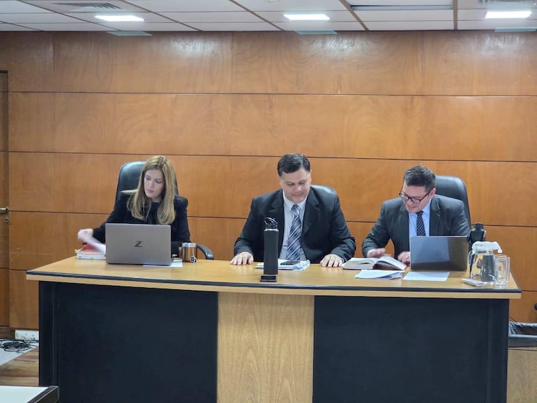 Inés Galarza sentada a la izquierda revisa documentos, mientras Juan Dávalos y Pablino Barreto están detrás de un escritorio revisando. Ambiente de oficina.
