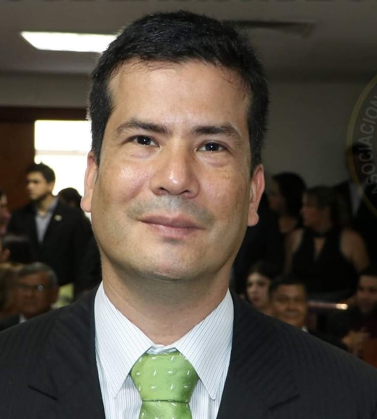 Hugo Alberto Oxilia Vera en primer plano, con traje negro, camisa blanca y corbata verde, en un ambiente formal con fondo desenfocado.