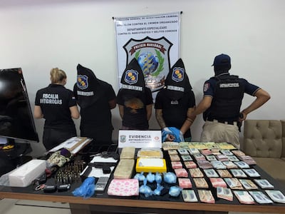 Cinco personas de espaldas, con camisetas negras y capuchas, frente a una mesa con paquetes y armas en ambiente tenso.