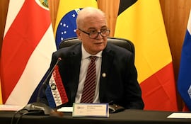 Dr. Emiliano Rolón Fernández sentado al micrófono, con traje oscuro y corbata rayada, frente a banderas de varios países.