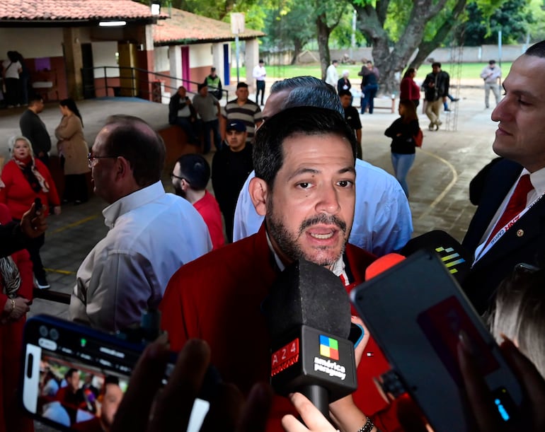 Raúl Latorre con chaqueta roja rodeado de micrófonos, mientras habla a una multitud en un entorno al aire libre.